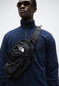 Μαύρο τσάντα χιαστί με υφή στην εξωτερική πλευρά, που διαθέτει ένα εξέχον λευκό λογότυπο. Ρυθμιζόμενος ιμάντας και ελαστικές λεπτομέρειες για ασφαλές κλείσιμο.