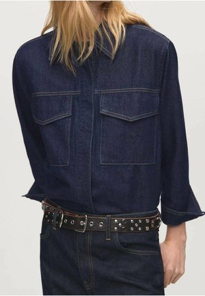 Personne portant une chemise en denim bleu foncé avec des poches à rabat et un pantalon en denim assorti, maintenu par une ceinture noire à clous argentés.