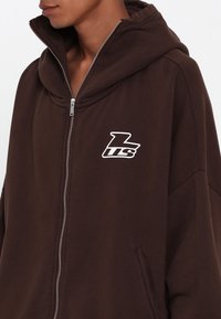 Sweat à capuche marron avec fermeture éclair, grande capuche, tissu texturé et poches latérales. Présente un logo blanc sur la poitrine pour la marque.
