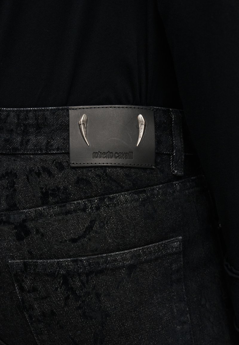 Primo piano di jeans in denim nero con una stampa leopardata discreta, caratterizzati da una patch in pelle nera con la scritta "roberto cavalli" e due borchie d'argento a forma di artiglio.