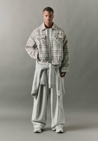 Šedá kostkovaná bunda s kapsami na hrudi, nošená přes světle šedý ribbed svetr, v kombinaci s volnými šedými tepláky a bílými teniskami.
