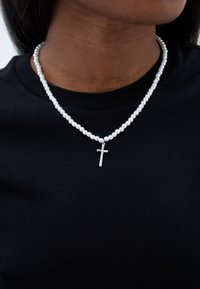 Vairakslāņu pērļu kaklarota ar mazām baltām pērļām un sudraba krusta piekariņu, uz melna auduma fona.