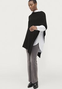Sort poncho-lignende top med batwing-ærmer over en hvid skjorte med krave, parret med grå vide jeans og sorte ankelstøvler. Simpelt design.