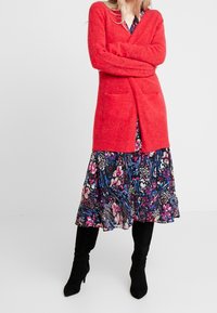 Röd, lång stickad cardigan med två framfickor, bärs över en svart blommig klänning med livfulla blå och rosa mönster. Svarta knästövlar.