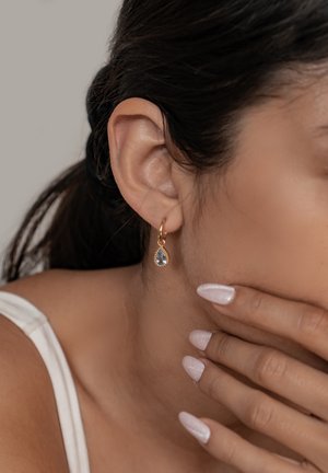 Gold hoop earring with a teardrop-shaped blue stone, worn on a pierced ear. Smooth skin and light pink nails are visible in the foreground.