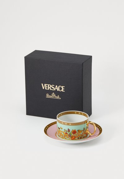 Keramiktässchen für Espresso mit türkisfarbenem Blumenmuster, goldenen Akzenten und pinkem Unterteller. Schwarze Schachtel mit der Aufschrift "Versace". Glatte Textur und abgerundete Form.