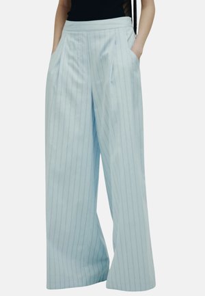 Person, die hellblaue, hoch taillierte, weit geschnittene Hose mit dünnen vertikalen Streifen und Seitentaschen trägt, kombiniert mit einem schwarzen Oberteil.