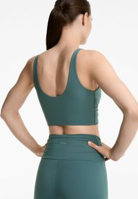Zöldes-kék sportos crop top széles pántokkal, sima textúrával és ráncolt részletekkel a háton, magasderekú leggingsszel párosítva.