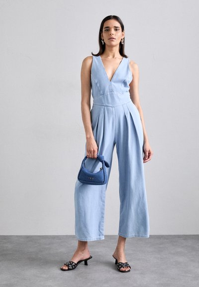 Jumpsuit en denim bleu clair avec un décolleté en V profond, des jambes larges et des plis. Le modèle tient un petit sac à main bleu marine et porte des escarpins noirs.