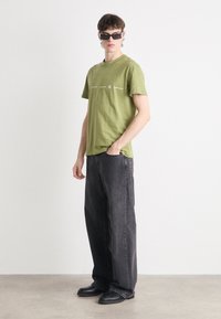 Camiseta de algodón verde con gráfico de texto blanco, combinada con jeans de mezclilla negros de corte amplio y zapatos negros. Los accesorios incluyen gafas de sol negras.