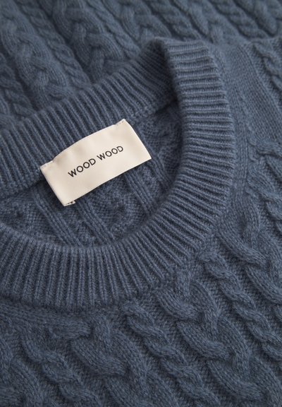 Svetr s kabelovým pletením v šedoblé barvě s žebrovaným kulatým výstřihem. Obsahuje tkanou texturu a látkovou nášivku s nápisem "WOOD WOOD."