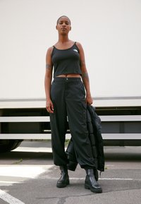 Woman stands outdoors wearing black crop top, loose cargo pants, chunky boots, holding a black jacket, with tattoos visible on her arms.