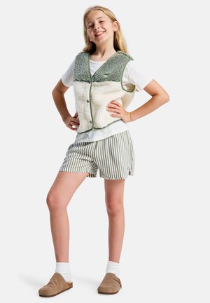 Gilet duveteux de couleur crème avec des accents verts, porté avec un T-shirt blanc et un short à rayures vertes. Des sabots marron et des chaussettes blanches complètent la tenue.