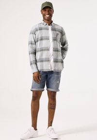 Camisa de cuadros de manga larga en gris y verde, llevada sobre una camiseta blanca. Pantalones cortos de denim azul con los puños doblados y zapatillas blancas. Gorra verde.
