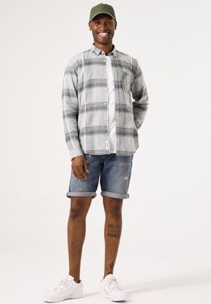 Long-sleeve plaid shirt in grey and green, worn over a white t-shirt. Blue denim shorts with rolled cuffs and white sneakers. Green cap.