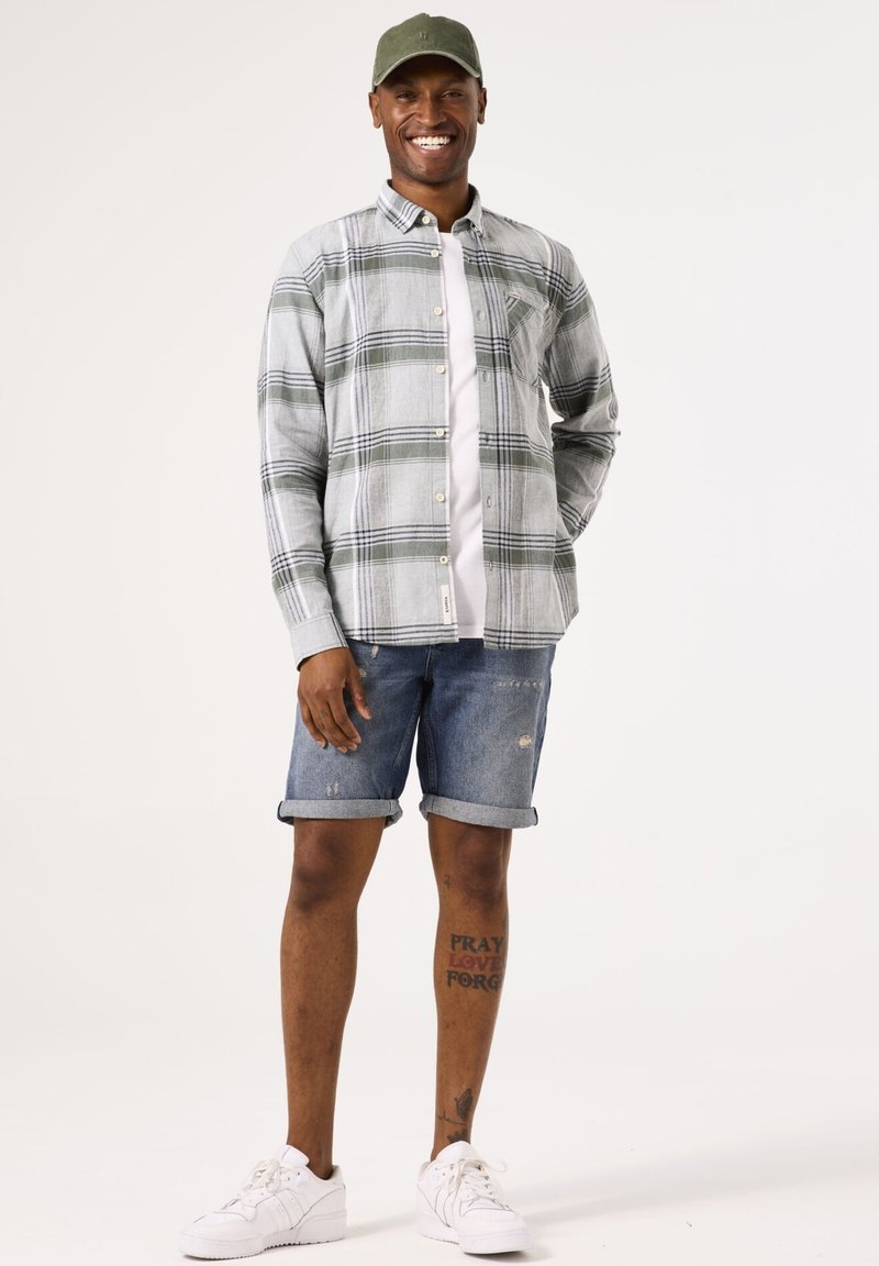Camisa de cuadros de manga larga en gris y verde, llevada sobre una camiseta blanca. Pantalones cortos de denim azul con los puños doblados y zapatillas blancas. Gorra verde.