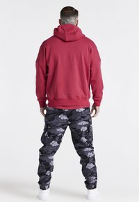 Felpa rossa con cappuccio dal taglio ampio, abbinata a pantaloni cargo neri con motivo mimetico e nuvole grigio chiaro. Sneakers bianche completano l'outfit.