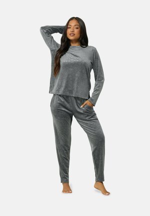 Femme debout pieds nus, une main derrière la tête, portant un ensemble assorti de vêtements d'intérieur gris doux avec un haut à manches longues et un pantalon.