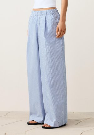 Žena nosi široke plave i bijele prugaste hlače s elastičnim strukom, uparene s crnim sandalama i bijelim kratkim topom.