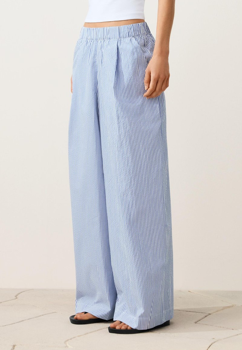 Femme portant un pantalon large rayé bleu et blanc avec une taille élastique, associé à des sandales noires et un top court blanc.