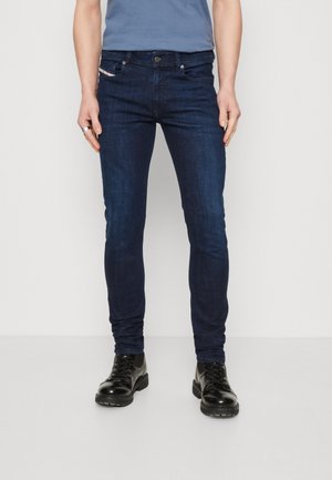 Personne portant un jean slim bleu foncé, des chaussures en cuir noires à lacets et une chemise bleu à manches courtes, debout devant un fond blanc uni.