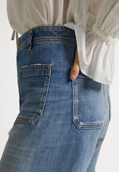 Άτομο που φοράει μπλε τζιν τζιν με το χέρι εν μέρει στην πίσω τσέπη, συνδυασμένο με λευκό διάφανο πουκάμισο με μακριά μανίκια.