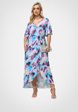 Kvinde iført en blå og lilla mønstret omslagskjole med flagrende ærmer, der holder en sølvfarvet clutch og har sølvfarvede hæle på.