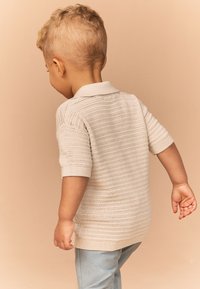 Lysebeige strikket polo med korte ærmer, der har en striped tekstur. Lyseblå bukser fuldender outfittet. Barn vist bagfra.