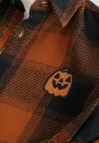 Zwarte en oranje geruite blouse met een kleine geborduurde glimlachende jack-o'-lantern pompoen bij de kraag.