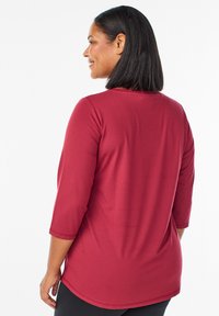 Burgundin värinen kolmannen neljänneksen hiha paita, joka on valmistettu sileästä kankaasta, siinä on pyöreä pääntie ja hieman kaareva helma rentoa istuvuutta varten.