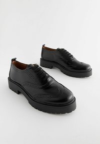 Zapatos de cuero negro con detalles perforados, punta redondeada y suela de goma gruesa. Presenta cordones y una pestaña en la parte posterior para facilitar su uso.
