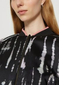 Veste bombardier noire avec un motif tie-dye en blanc et lavande. Elle présente une fermeture éclair et une bordure de col contrastante en rose. Texture lisse.