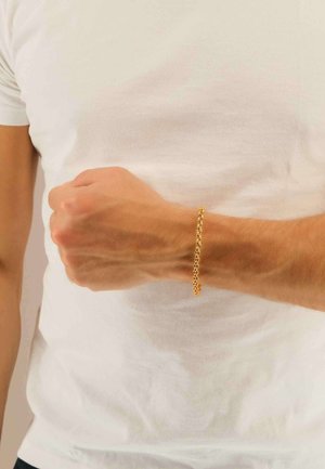 Braccialetto con catena d'oro al polso, con maglie incatenate e una texture liscia. Indossato con una t-shirt bianca semplice.