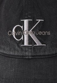 Black denim cap featuring embroidered logo with "CK" and "Calvin Klein Jeans" in gray. Textured fabric and curved brim design.