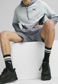 Personne assise sur un bloc blanc portant un hoodie Puma gris et blanc, un short assorti, des chaussettes Puma noires et des baskets noires.