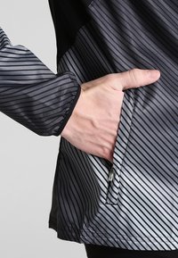 Giubbotto leggero con strisce diagonali nere e grigie, dotato di una tasca con zip sul lato e polsini elasticizzati. Tessuto liscio e texture.