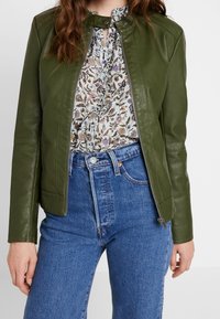 Veste en cuir verte au design ajusté, dotée d'un col haut et d'une fermeture éclair, associée à un blouse à motif floral et à un jean bleu taille haute.