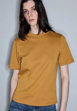 Młoda osoba z długimi ciemnymi włosami, ubrana w musztardowożółtą koszulkę z krótkim rękawem i niebieskie dżinsy, stojąca na tle jednolitego jasnego tła.