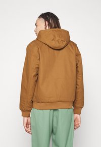 Brown hooded jacket with a relaxed fit, textured fabric, and ribbed cuffs. Worn over light green pants, showcasing a casual style.