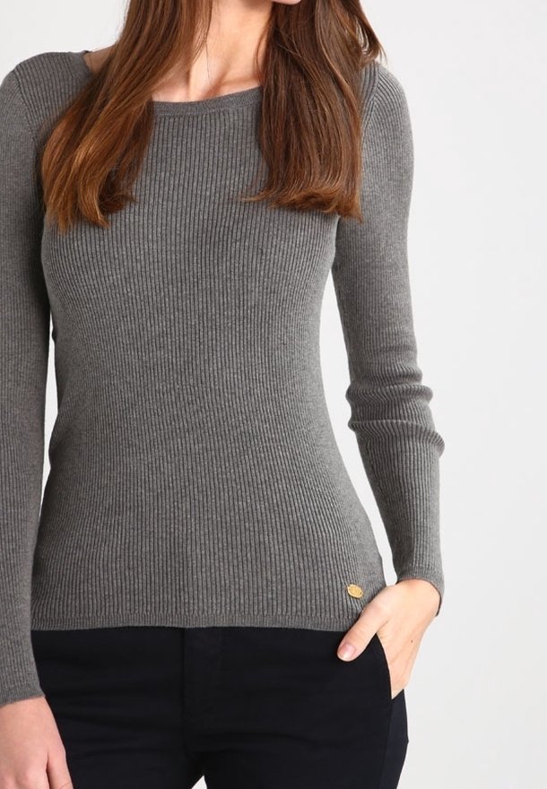Femme portant un pull gris moulant à côtes avec manches longues et col rond, associé à un pantalon noir, main dans la poche.