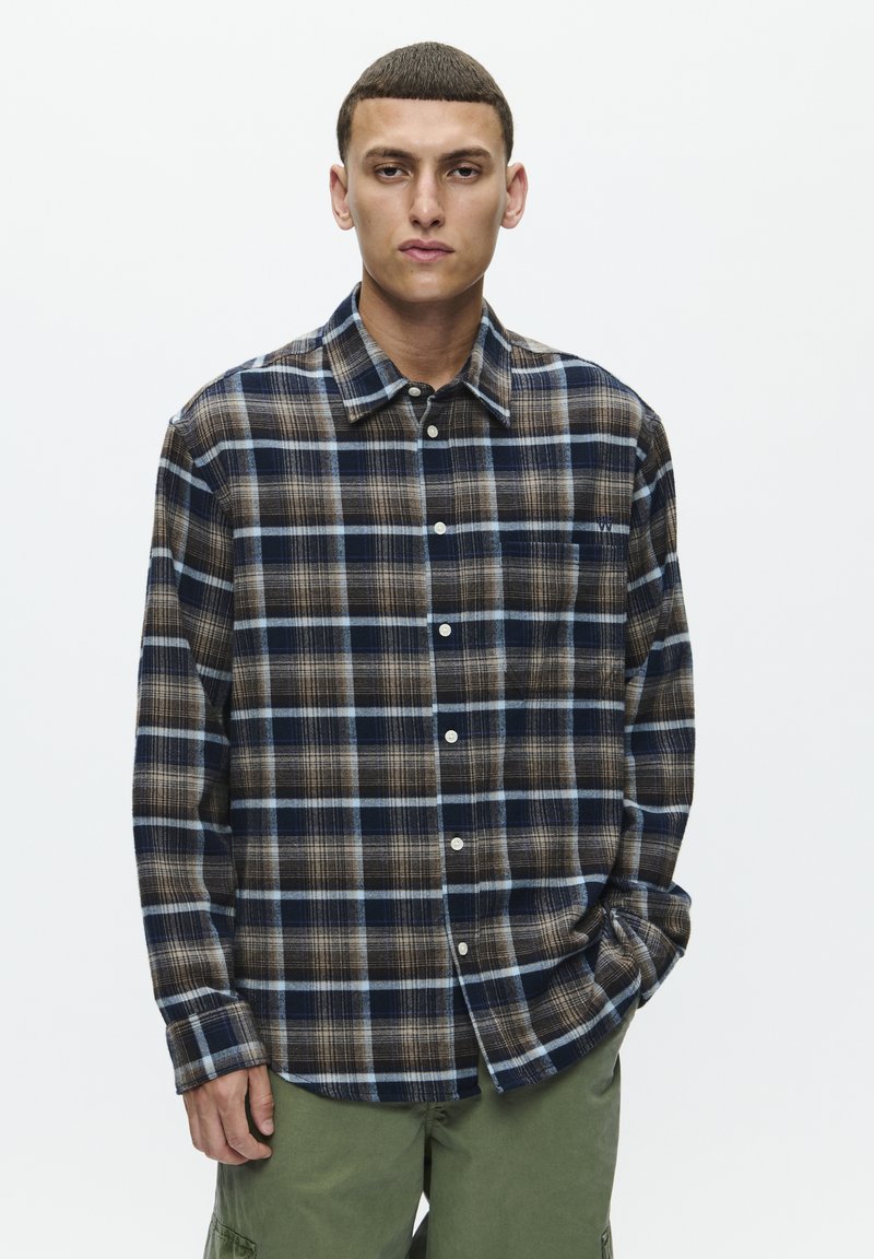 Flannel skjorte med et navyblåt, brunt og lyseblåt ternet mønster, med knaplukning og spidse kraver. Inkluderer hvide knapper.