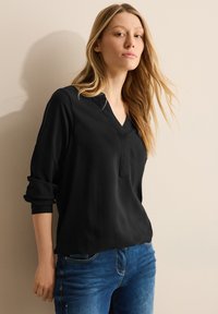 Zwarte, gestructureerde blouse met een diepe V-hals en omgeslagen mouwen, gecombineerd met blauwe jeans. De stof lijkt lichtgewicht en casual te zijn.