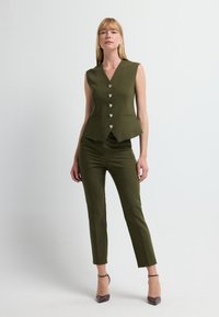 Oliwkowozielona kamizelka bez rękawów z dekoltem w serek, zapinana na pięć guzików, z dwoma przednimi kieszeniami, w zestawieniu z dopasowanymi spodniami i butami na obcasie.
