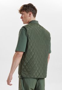 Zelená prošívaná vesta s diamondovým vzorem, s límcem a bez rukávů, nošená přes krátký rukáv zelené košile. Plochá a hladká textura.