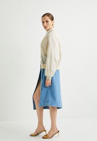 Chaqueta bomber transparente de color beige claro con mangas globo, combinada con una falda de mezclilla azul con una abertura alta en el muslo y tacones abiertos dorados.