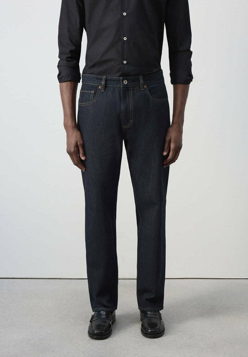 Homme portant un jean bleu foncé, une chemise noire boutonnée avec les manches retroussées jusqu'aux coudes, et des chaussures noires, debout devant un fond clair uni.