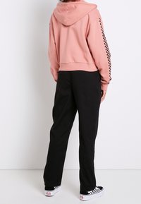 Persona che indossa un hoodie rosa con una striscia a scacchi neri sulla manica, pantaloni neri e scarpe slip-on a scacchi bianco e nero, in piedi con la schiena rivolta.
