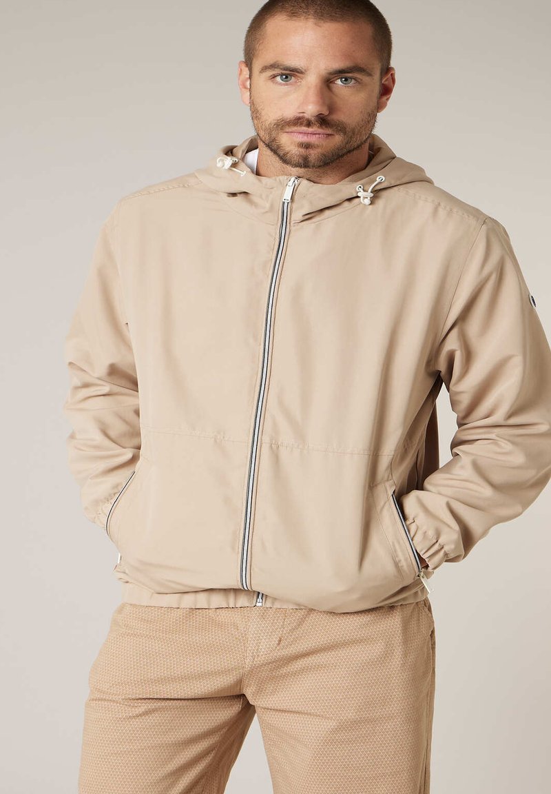 Homme portant une veste à capuche beige zippée avec les mains dans les poches et un pantalon beige clair à motif, debout devant un fond uni.