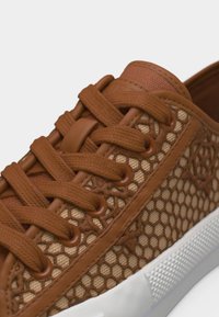 Καφέ αθλητικό παπούτσι με κορδόνια, σχέδιο από δίχτυ μέλισσας και λευκή υφή από καουτσούκ στη σόλα, κοντινή λήψη στα κορδόνια και το πλαϊνό σχέδιο.