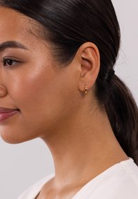 Kleine goldene Creolen mit verdrehter Textur. An dem Ohr getragen, zeigen sie ihre kreisförmige Form und die reflektierende Oberfläche.